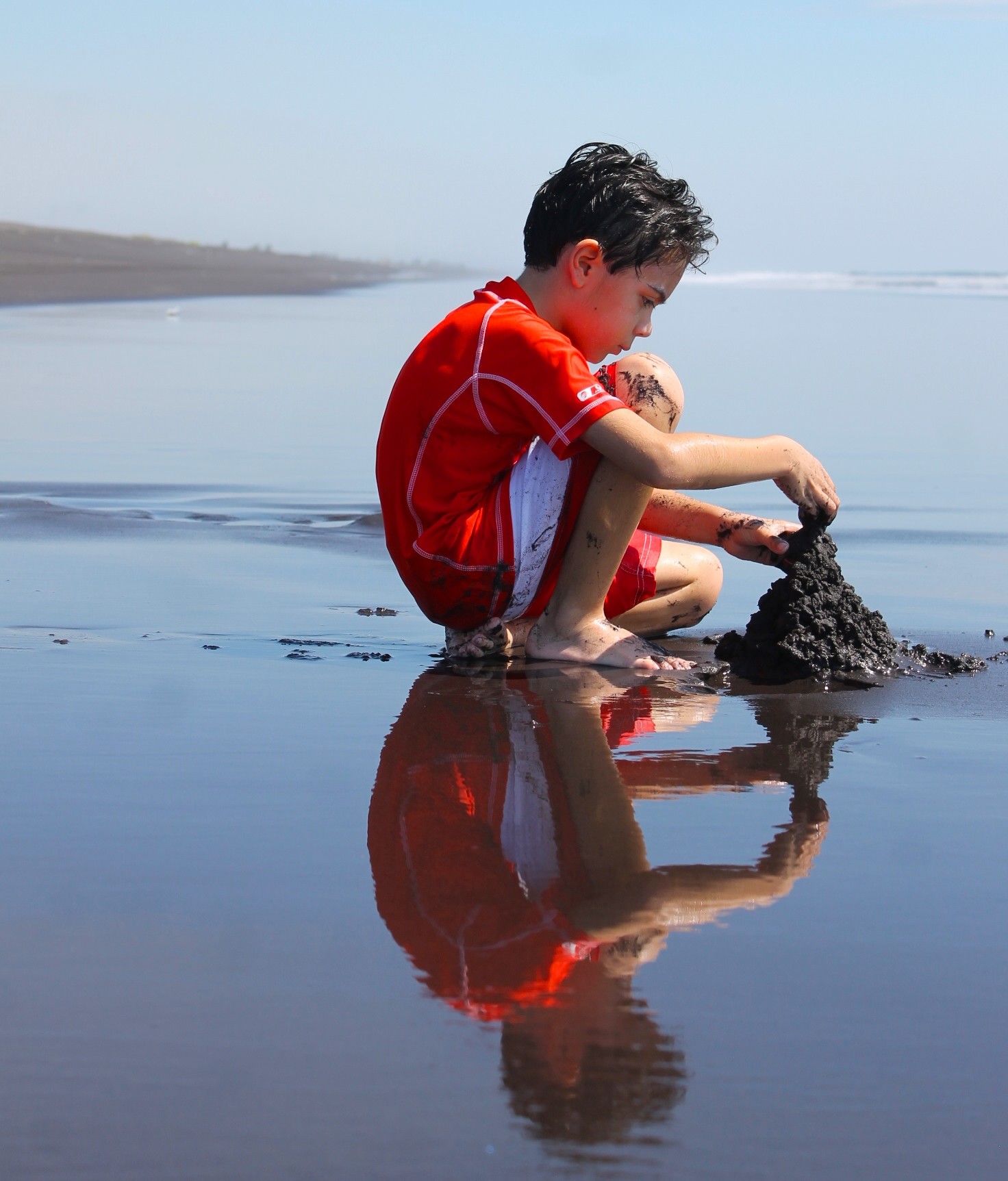boy playing with sand at black sand beach with reflection in the water
