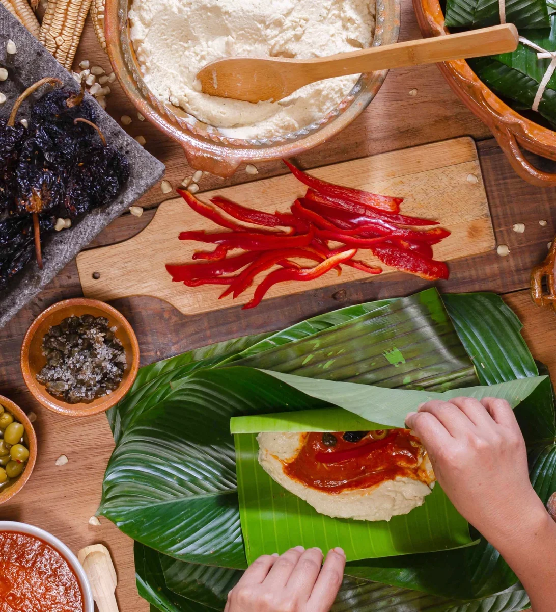 Manos doblando la hoja de banano sobre la masa y el recado, con chiles secos, aceitunas y alcaparras