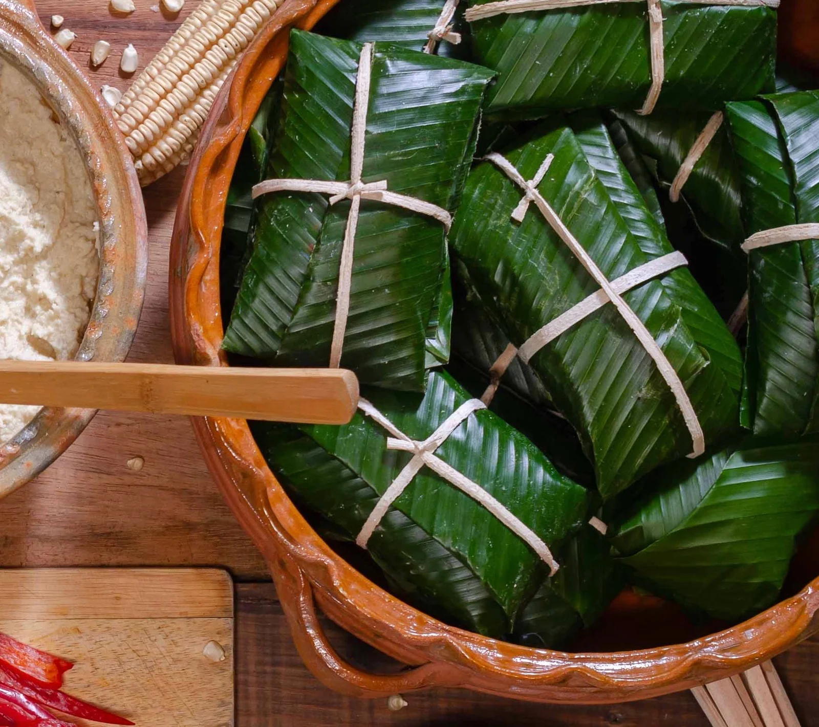Tamales en hoja de banano amarrados con cuerda, en una olla de barro junto a masa cocida y una mazorca