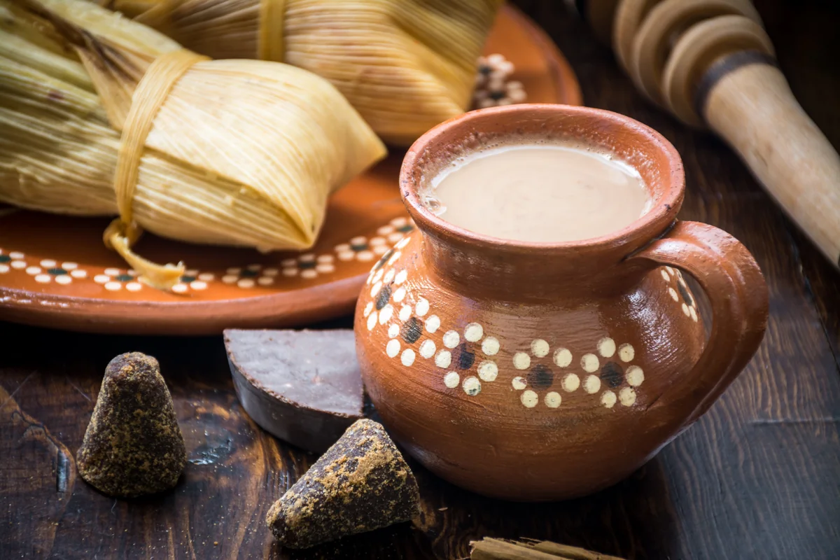 Tamales en tusa en un plato de barro con un jarrito de chocolate caliente, chocolate en tablilla y piloncillo