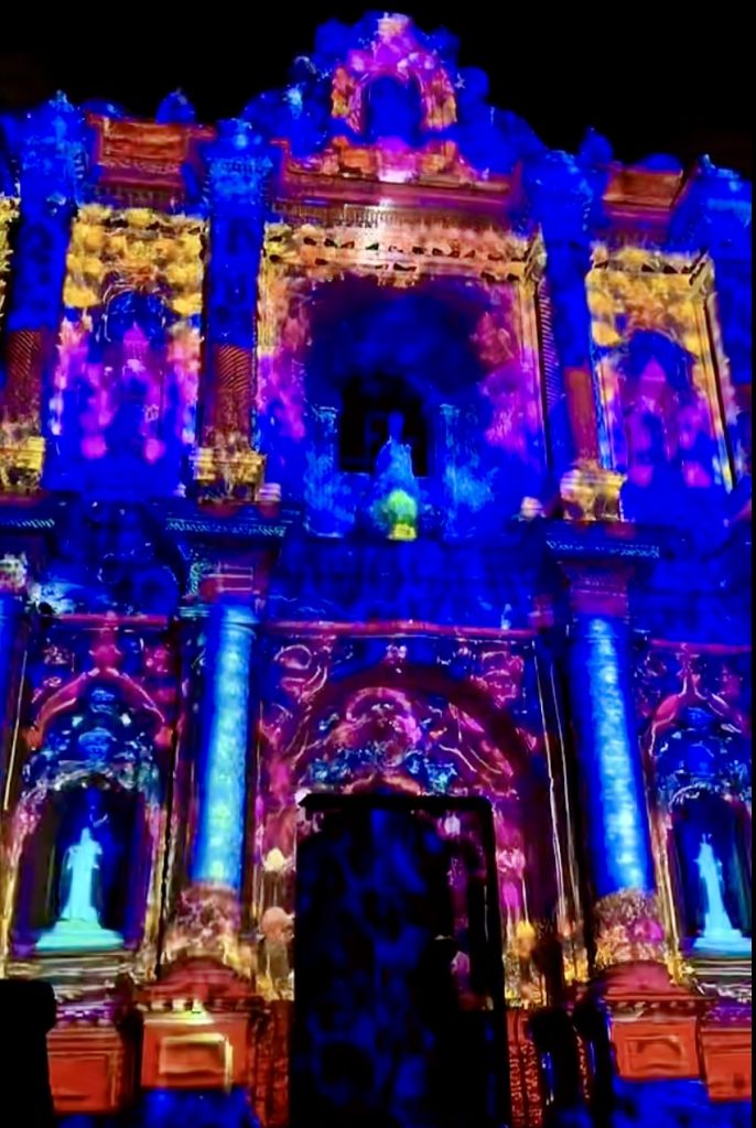 Illuminated colonial church facade with vibrant blue, purple, and yellow videomapping projections.