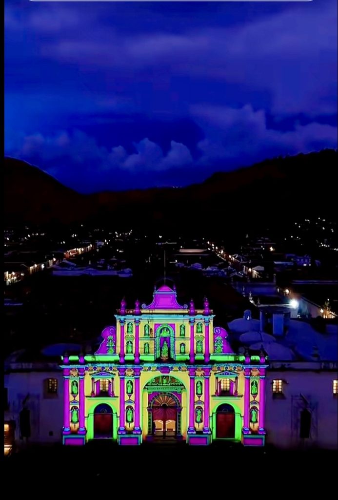 Bright pink and green videomapping lights cover the facade of a colonial building in Antigua, Guatemala at dusk.
