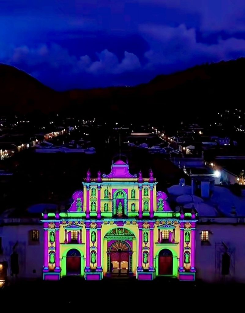 Bright pink and green videomapping lights cover the facade of a colonial building in Antigua, Guatemala at dusk.