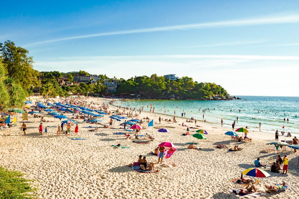 Crowded tropical beach scene at Kata Beach in Phuket, Thailand, with colorful umbrellas, sunbathers, and swimmers in turquoise water.