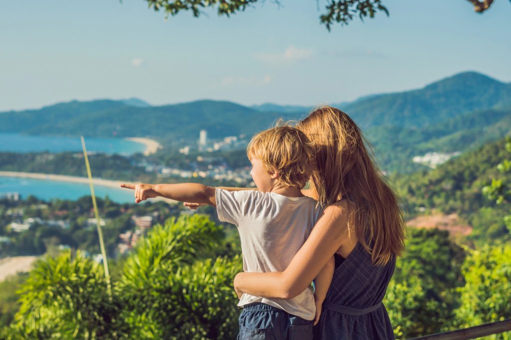 Mother holding her young son while he points toward the coastline from Karon Viewpoint in Phuket, with ocean and lush mountains in the background.
