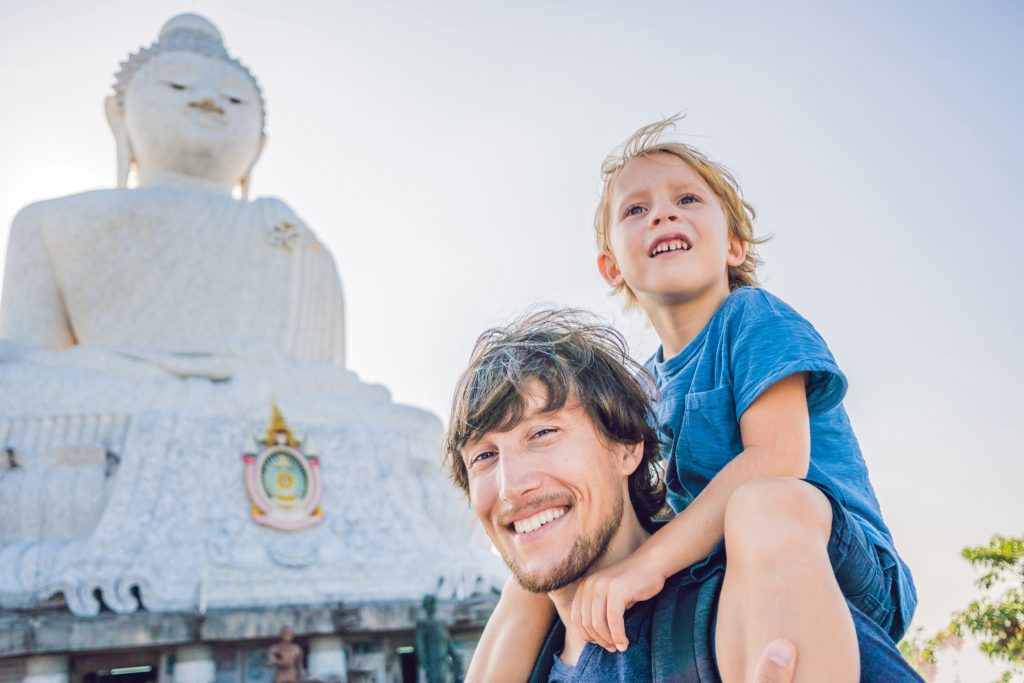 Father carrying his son on his shoulders in front of the Big Buddha statue in Phuket, Thailand, under clear skies.