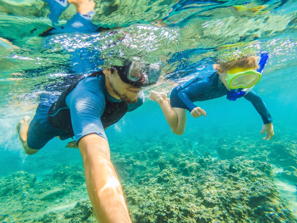 Underwater view of a father and child snorkeling in crystal-clear tropical water above coral reefs in Thailand.