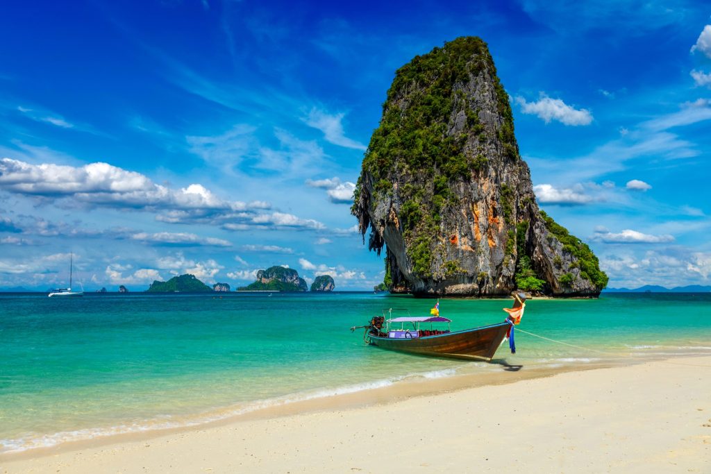 Traditional Thai longtail boat anchored near a dramatic limestone karst island with turquoise water and blue skies.