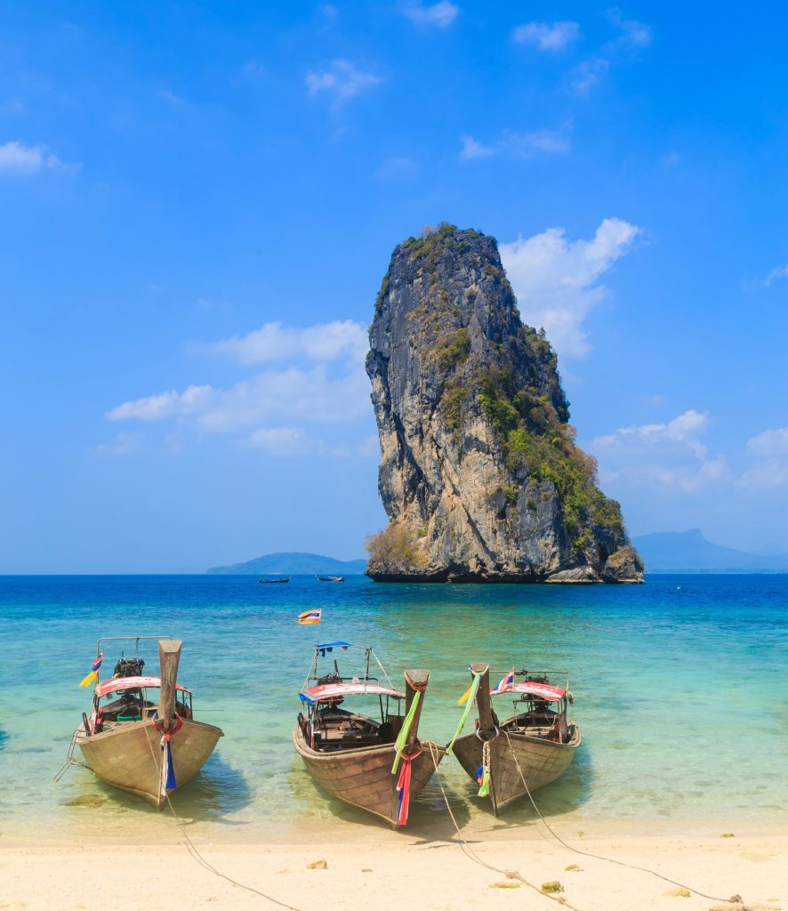 Three longtail boats anchored on clear, shallow waters at Ao Nang Beach with a dramatic limestone island off the Krabi coastline.