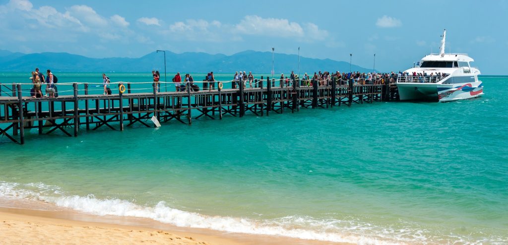 Long pier with people boarding the Lomprayah high-speed catamaran ferry in turquoise waters off Koh Samui, Thailand.