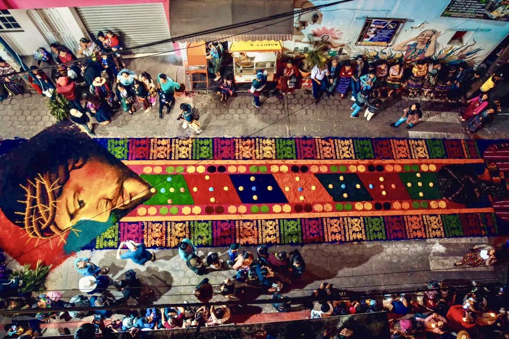 Stunning aerial view of an elaborate colored sawdust alfombra depicting the face of Christ surrounded by traditional Maya textile patterns on the streets of San Juan La Laguna during Holy Week — read our complete guide to Semana Santa at Lake Atitlán Guatemala and what happens in every village