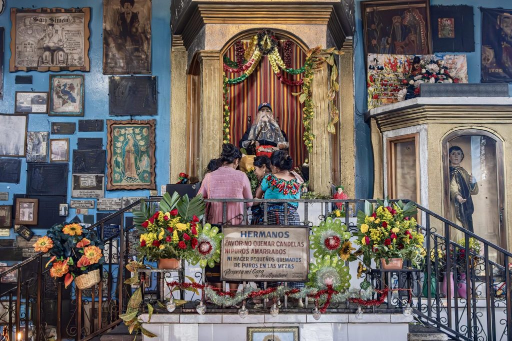 Interior of the San Simón chapel in San Andrés Itzapa with devotees making offerings, retablo paintings on the blue walls, and the San Simón figure in his decorated niche — Maximón San Andrés Itzapa, Guatemala
