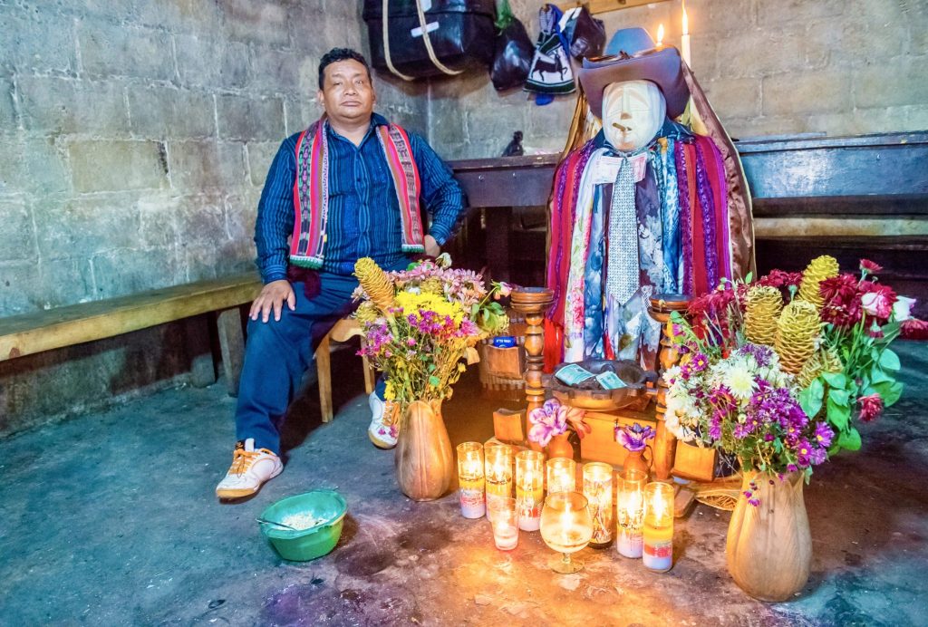 A cofrade seated beside Maximón surrounded by candles and flower offerings in a dimly lit room — visiting Maximón in Guatemala, Santiago Atitlán