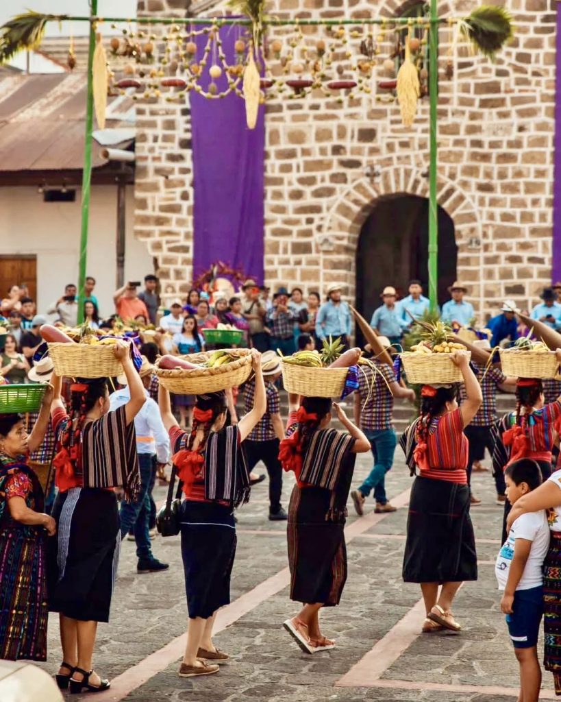 Indigenous women in traditional San Juan La Laguna huipiles carrying large wicker baskets of tropical fruit on their heads during the traída de fruta Holy Wednesday procession in front of the church — read our complete guide to Holy Week at Lake Atitlán Guatemala and Semana Santa traditions in every village