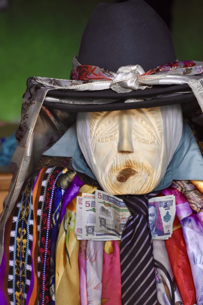 Close-up of Maximón's carved wooden face with a cigar between his lips and Guatemalan banknotes tucked into his scarves — visiting Maximón in Santiago Atitlán, Guatemala