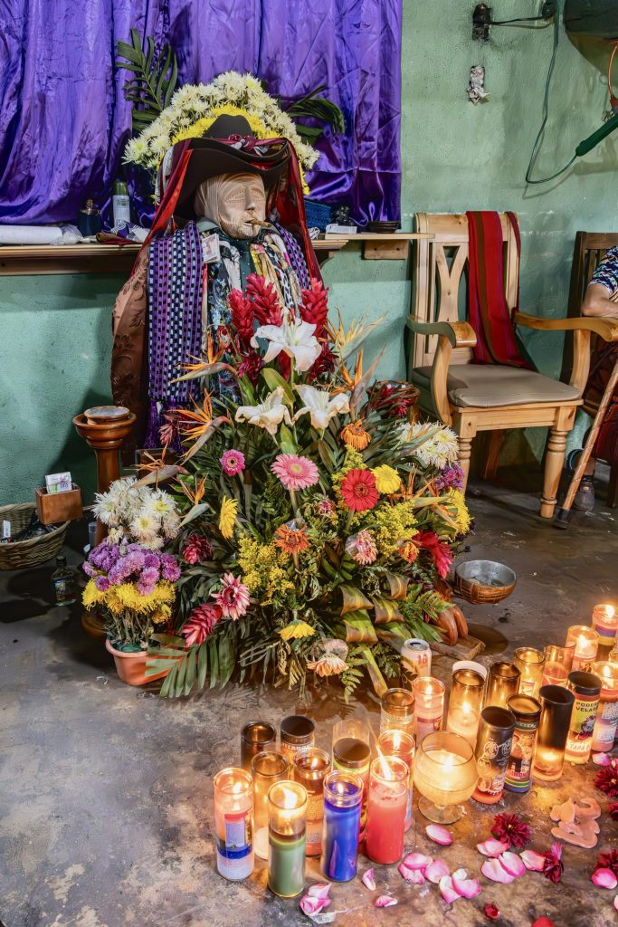 Maximón seated in his chapel surrounded by tropical flower offerings and rows of burning candles — Maximón Santiago Atitlán, Guatemala