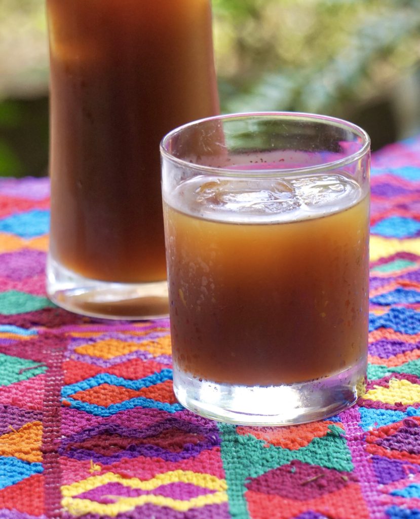 Glass of fresco de chilacayote over ice on a colorful Guatemalan textile with a pitcher in the background