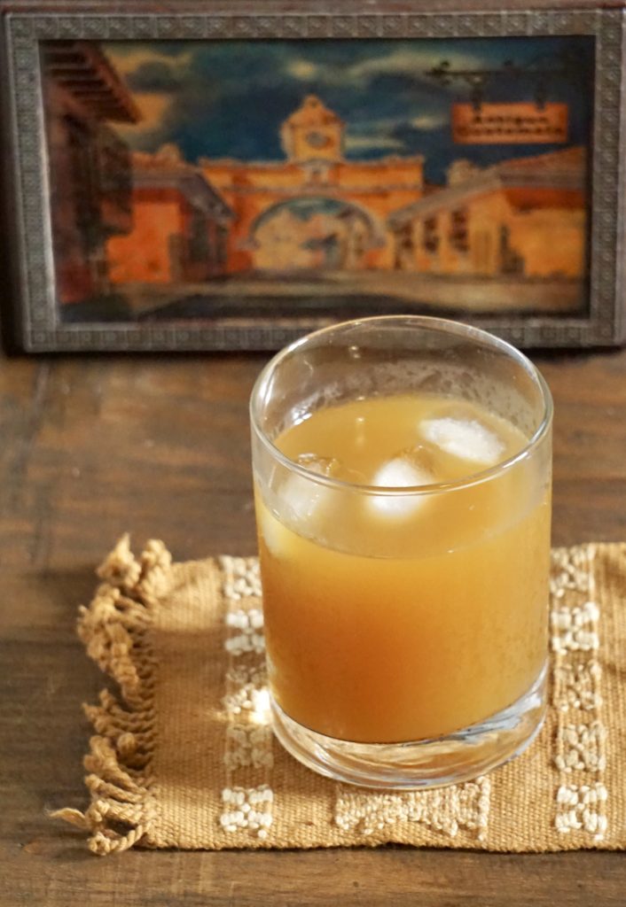 Fresco de suchiles served over ice in a glass with a painting of the Antigua Guatemala arch in the background