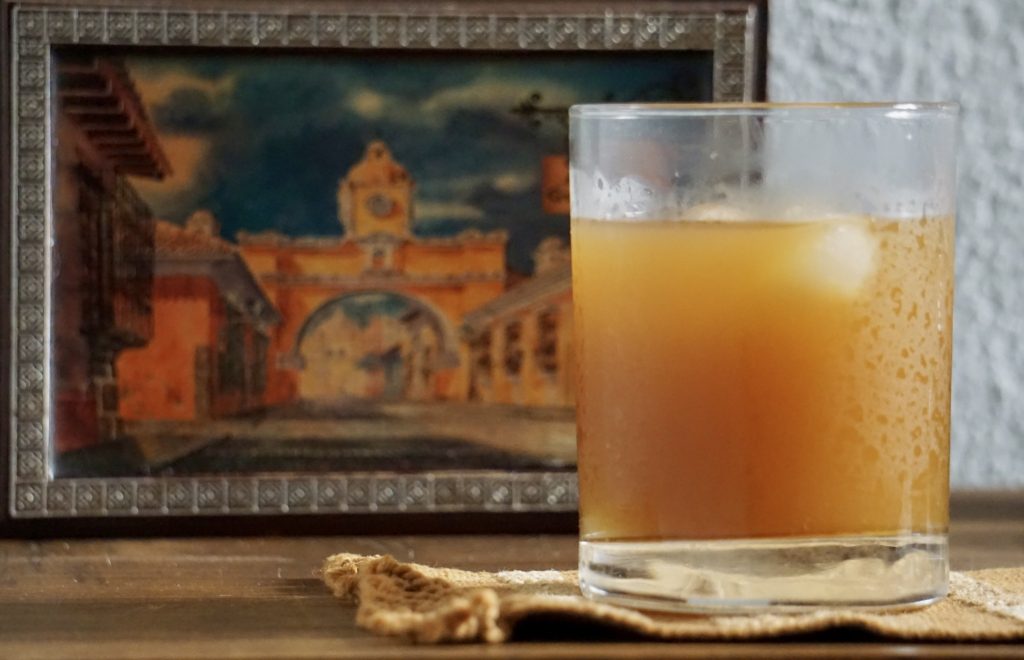 Receta de fresco de suchiles — vaso con fresco sobre tela de maguey con pintura de la Antigua Guatemala al fondo