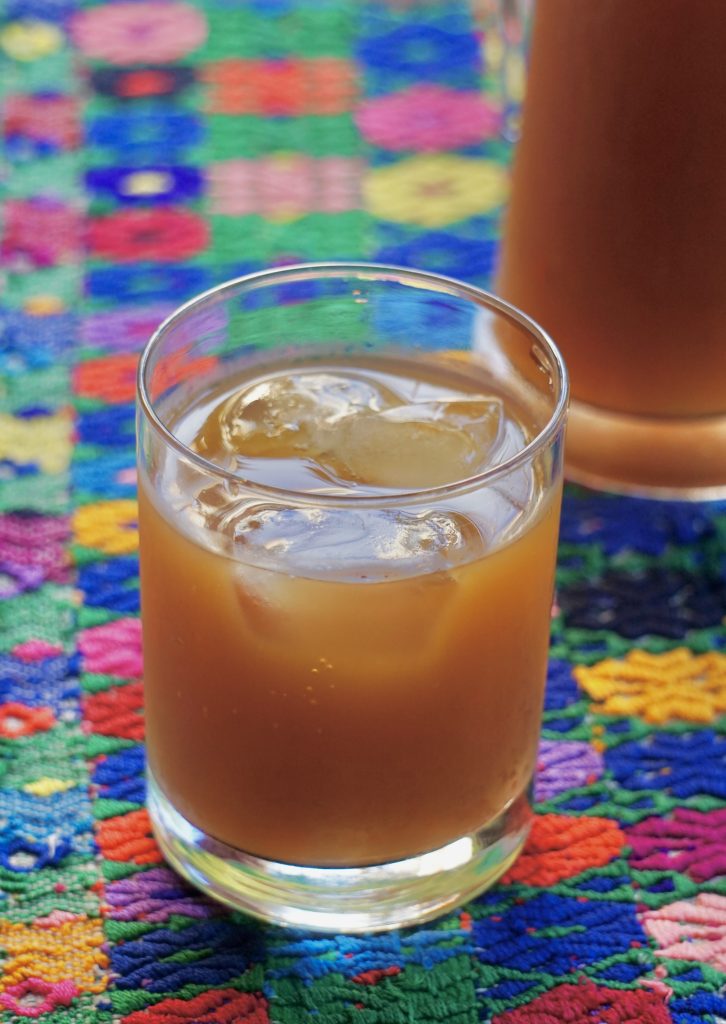 Glass of fresco de suchiles over ice on a colorful Guatemalan huipil textile with a glass pitcher in the background
