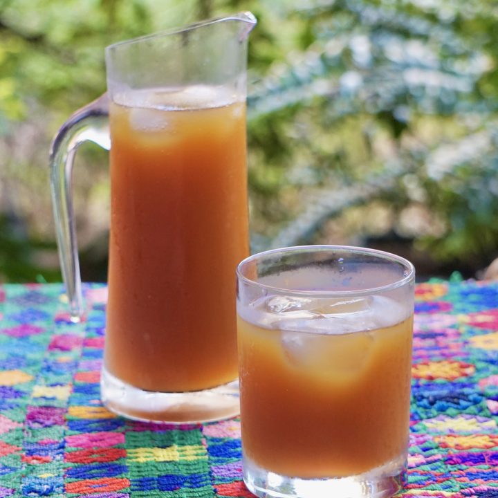 Pitcher and glass of fresco de suchiles over ice on a colorful Guatemalan textile with green garden in the background