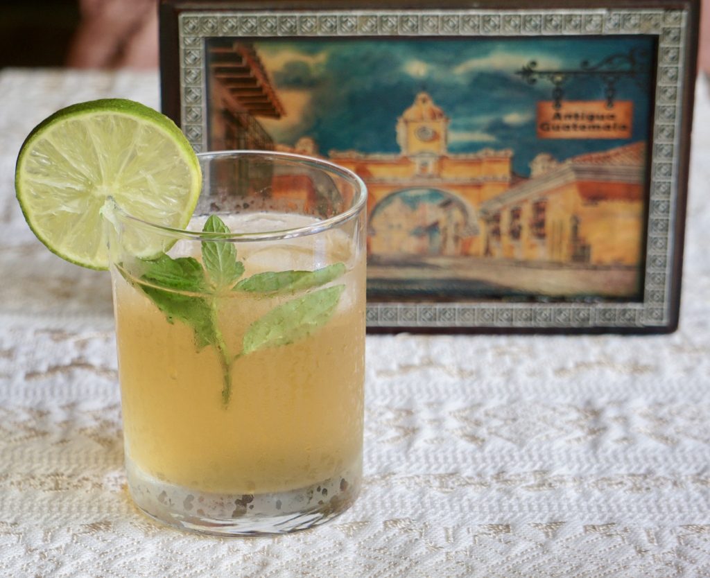 Vaso de fresco de chinchivir con hielo, limón y hierbabuena sobre mantel blanco con una pintura del Arco de Antigua Guatemala al fondo