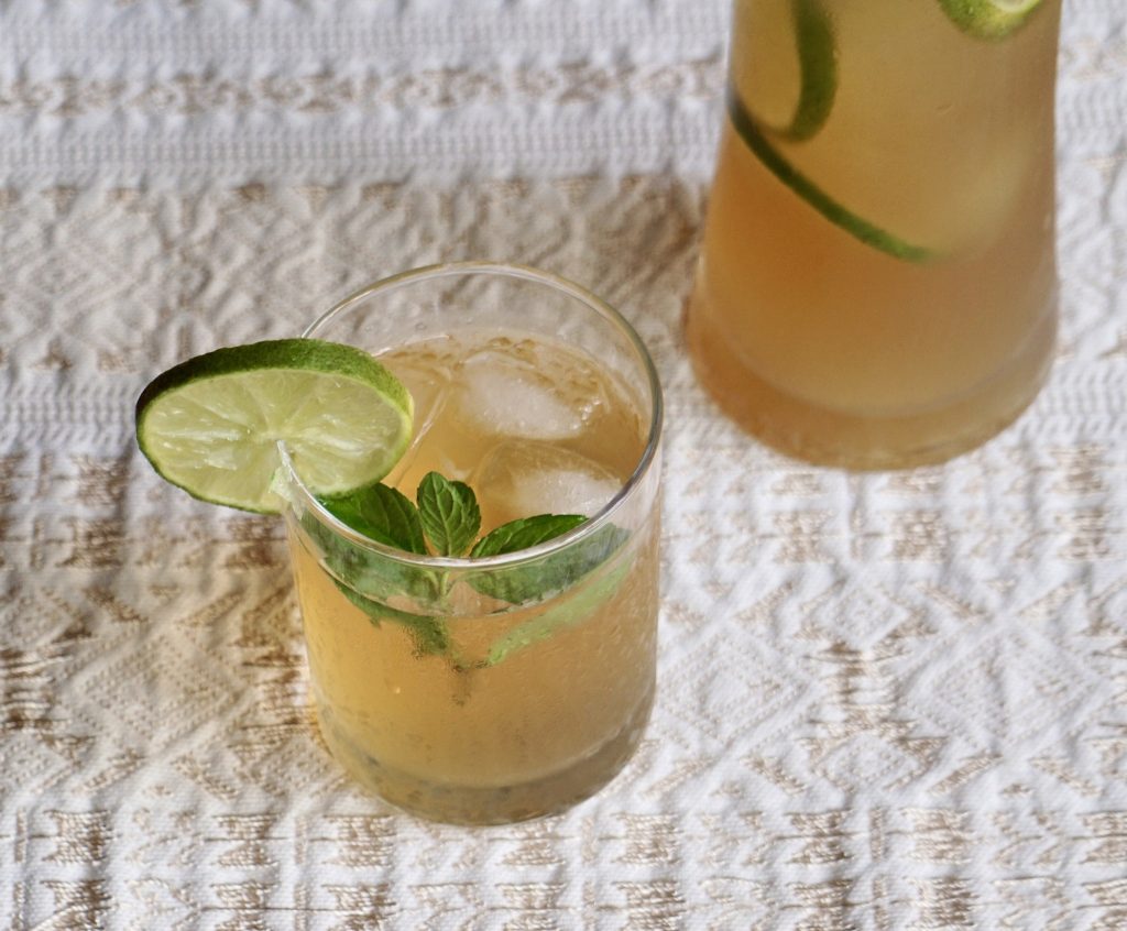 Glass of chinchivir over ice with lime and mint garnish next to a pitcher of the same drink on a white textured tablecloth