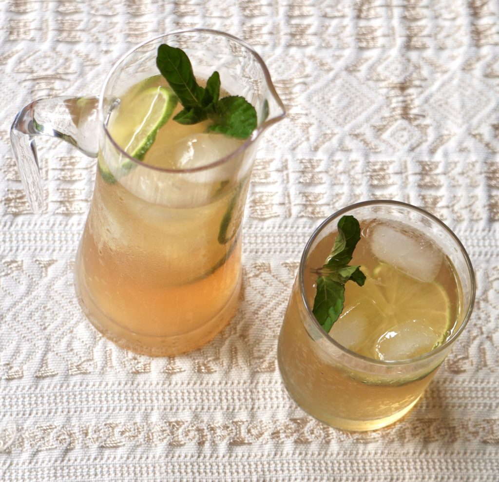 Vista superior de jarra y vaso de fresco de chinchivir con hielo, rodajas de limón persa y hojas de hierbabuena sobre mantel blanco