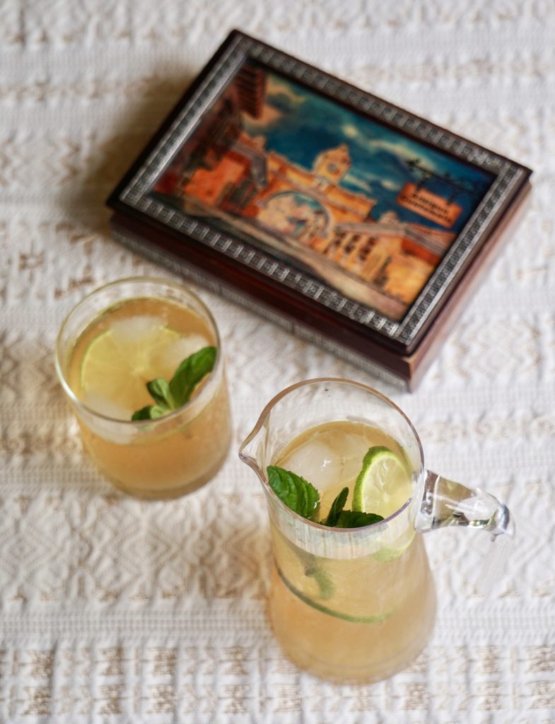 Overhead view of a glass and small pitcher of fresco de chinchivir with lime and mint on a white tablecloth next to a decorative Antigua Guatemala painting box