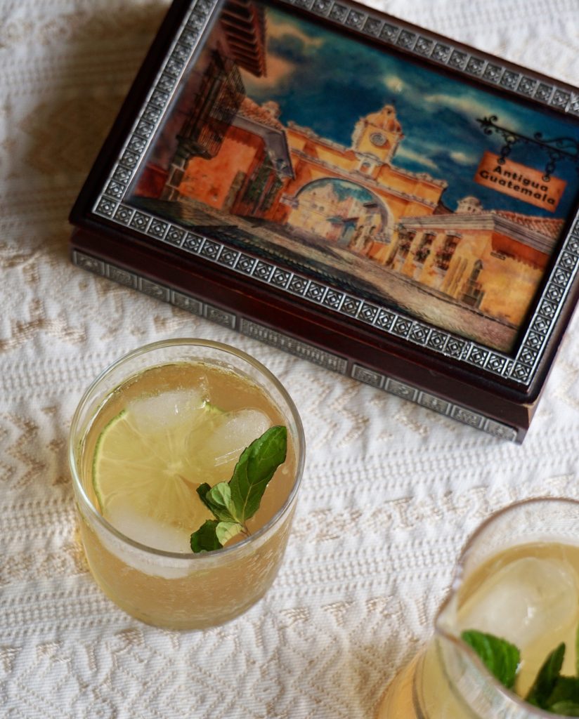 Vista superior de un vaso de fresco de chinchivir con limón persa y hierbabuena sobre mantel blanco con una caja decorativa de Antigua Guatemala al fondo