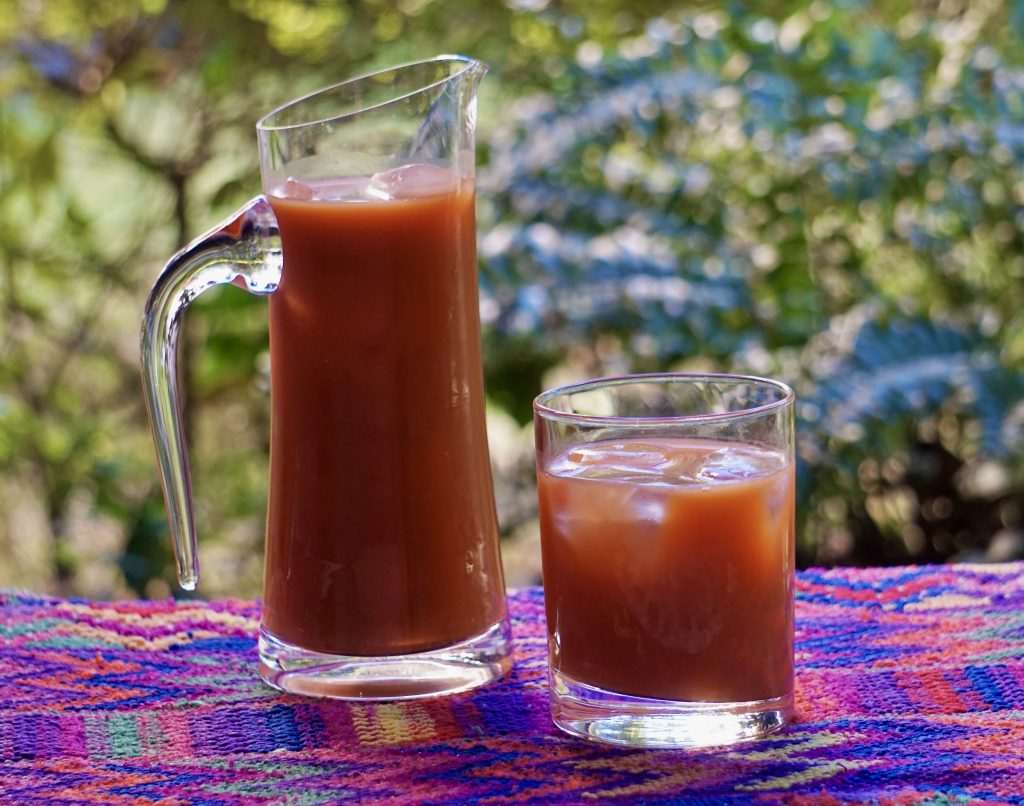 Pitcher and glass of fresco de tiste over ice on a colorful Guatemalan textile with a lush green garden in the background