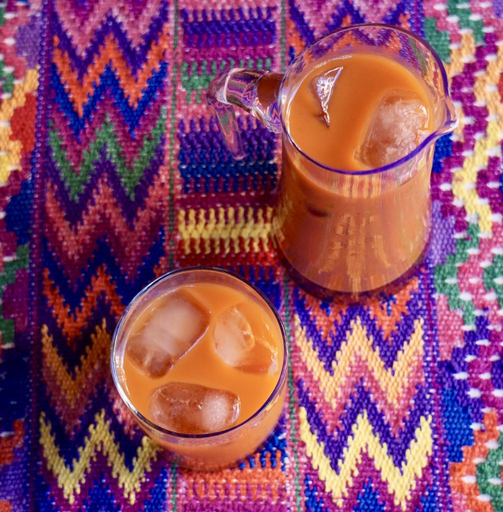 Vista superior de un vaso y una jarra pequeña de fresco de tiste con hielo sobre un mantel bordado guatemalteco de colores vivos