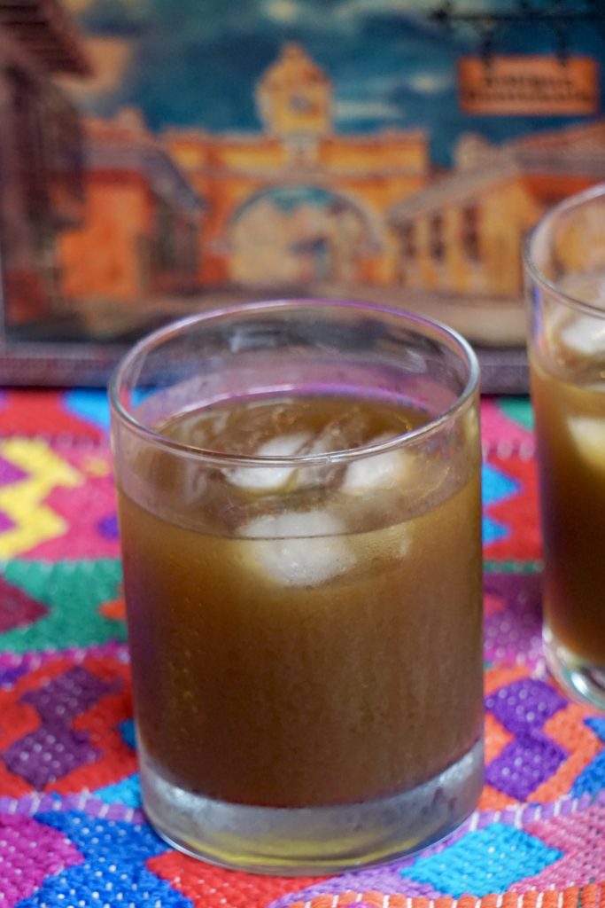 Two glasses of fresco de chilacayote over ice from above on a colorful Guatemalan textile next to a vintage Antigua Guatemala painting