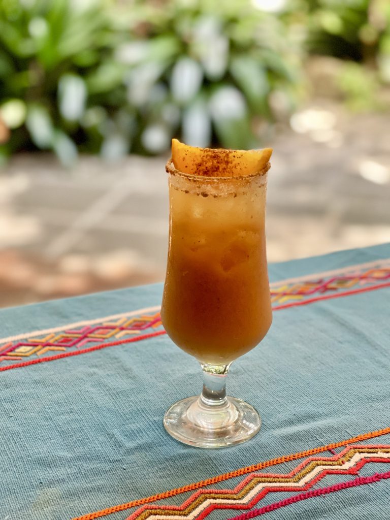 Glass of fresco de pepita with a spiced rim and mango garnish on a Guatemalan embroidered textile with a garden in the background