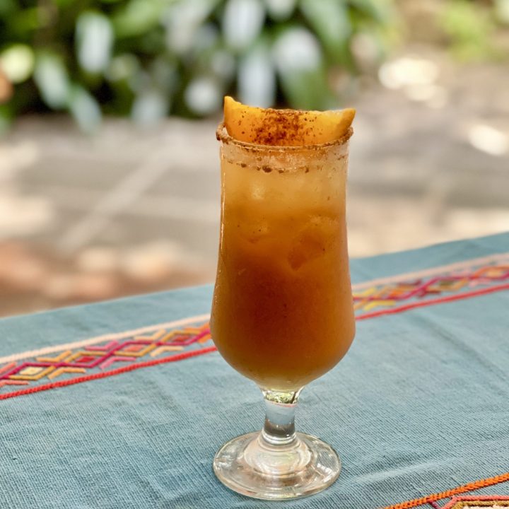 Glass of fresco de pepita with a spiced rim and mango garnish on a Guatemalan embroidered textile with a garden in the background