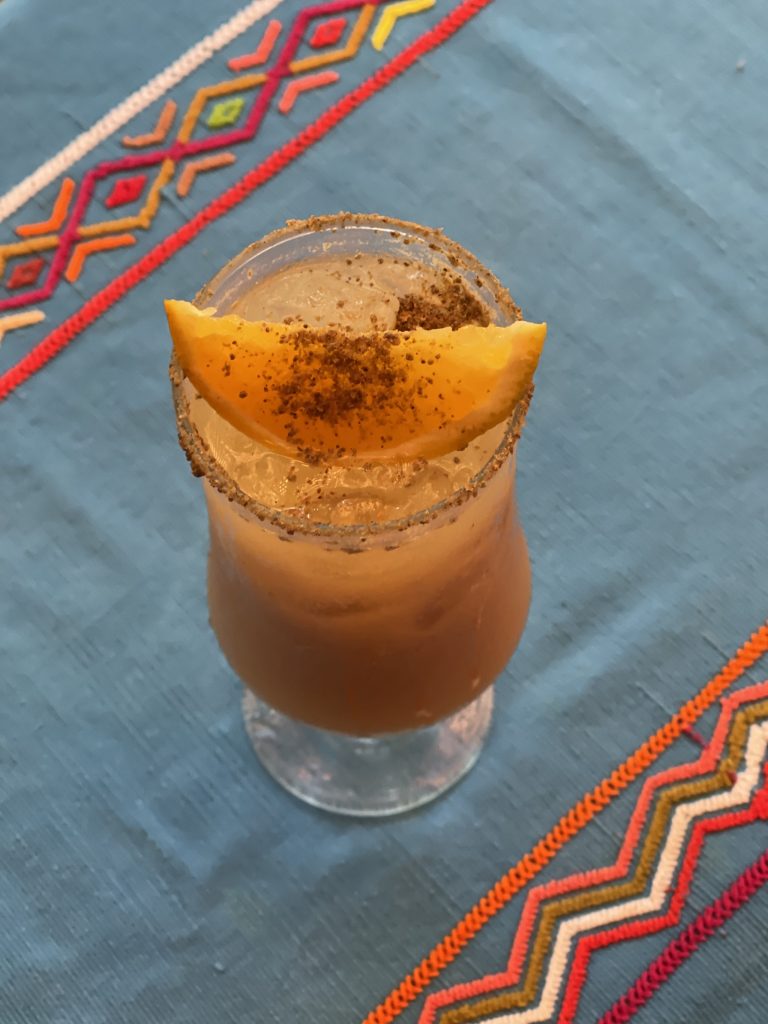 Overhead view of a fresco de pepita in a tall glass with a spiced pepita rim and cinnamon-dusted mango on a blue Guatemalan tablecloth