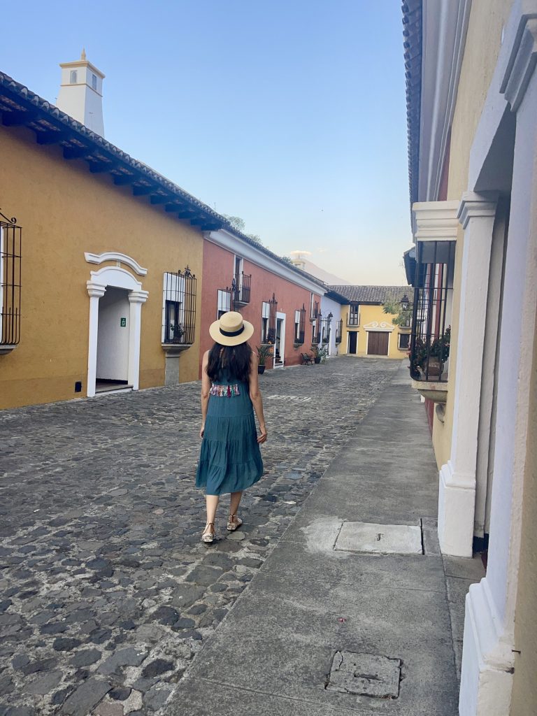 The private internal cobblestone street at Villa Colonial hotel in Antigua Guatemala, lined with colonial buildings in ochre and terracotta, with Volcán de Agua visible at the end