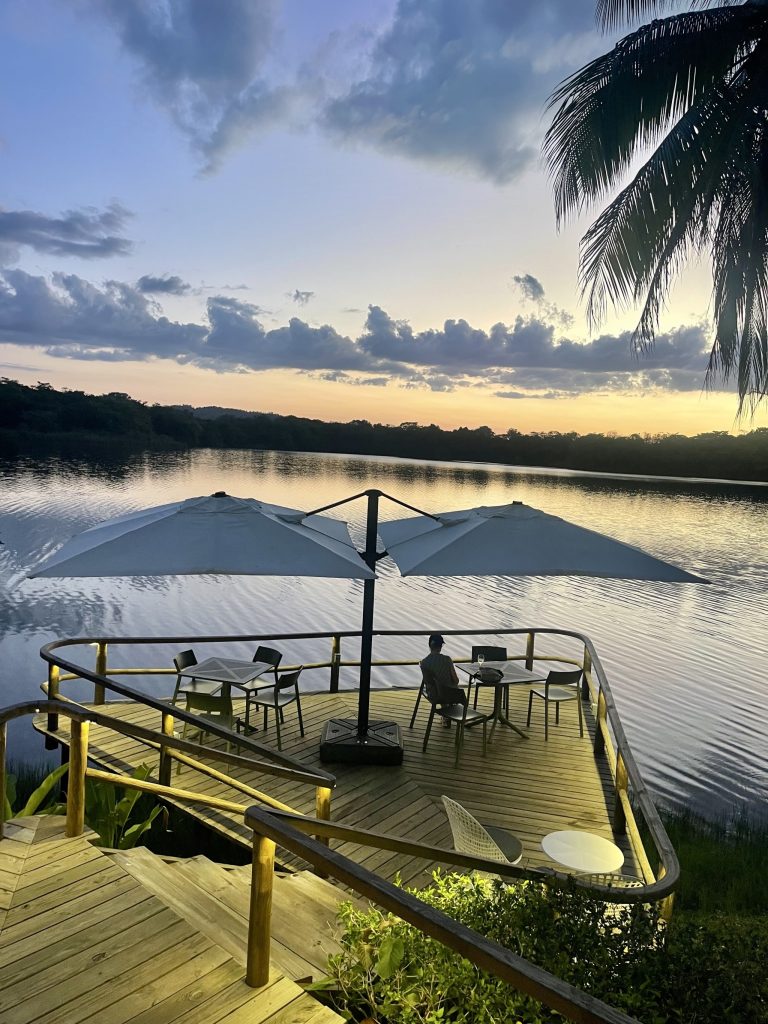 Terraza de madera del restaurante del Hotel Villa Maya en Petén Guatemala al atardecer, con mesas bajo sombrillas blancas sobre la laguna Petenchel, la silueta de una palmera y un cielo dramático en tonos naranja y morado reflejado en el agua tranquila 