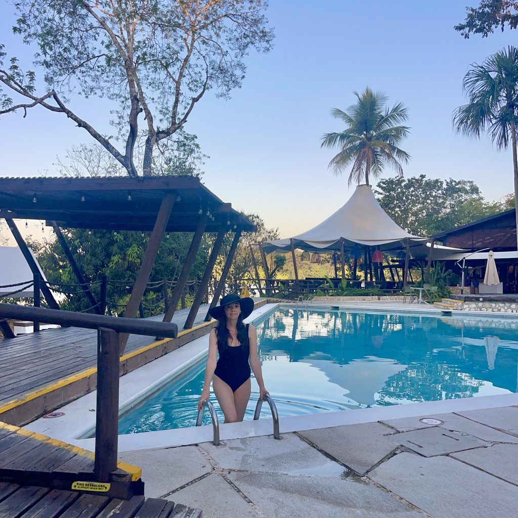 La piscina principal del Hotel Villa Maya en Petén Guatemala al atardecer, con el cielo anaranjado reflejado en el agua, palmeras silueteadas contra la luz y las lámparas de colores del restaurante iluminando el fondo