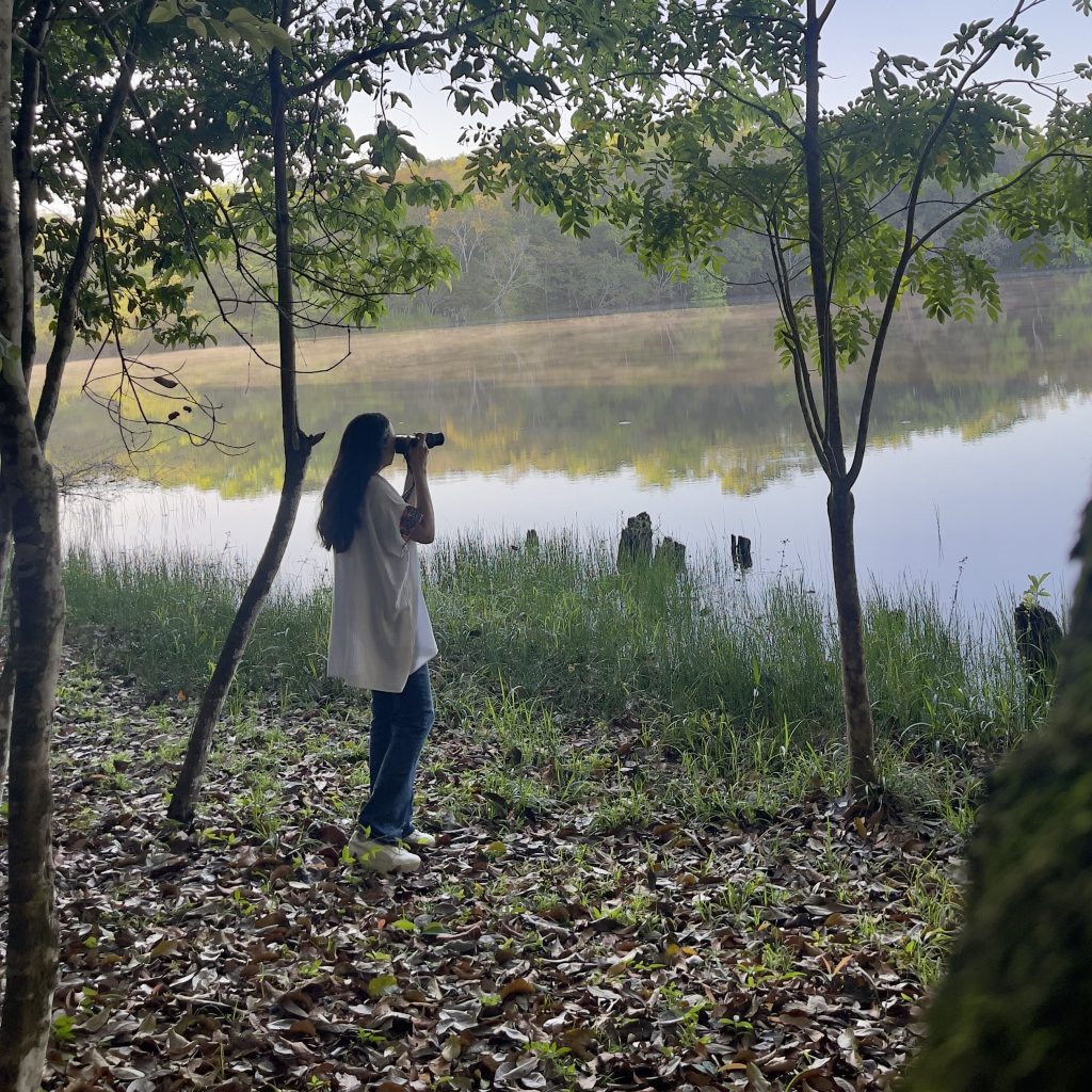 Person using binoculars to watch birds at the edge of the Petenchel lagoon on the nature trails at Villa Maya hotel in Petén Guatemala, with morning mist over calm water and dense jungle on the opposite shore