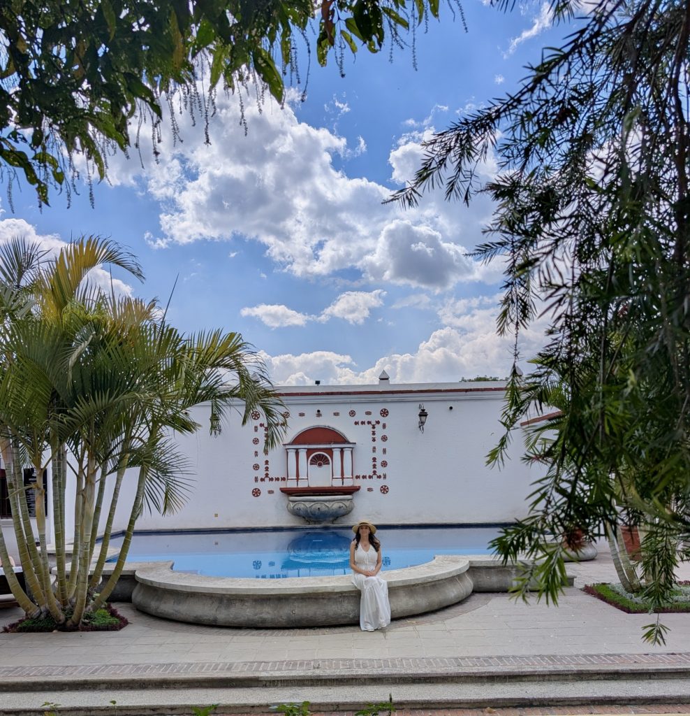 Mujer sentada en el borde de la piscina de Villa Colonial en Antigua Guatemala, rodeada de palmeras bajo un cielo azul con nubes, con la pared ornamental colonial de fondo