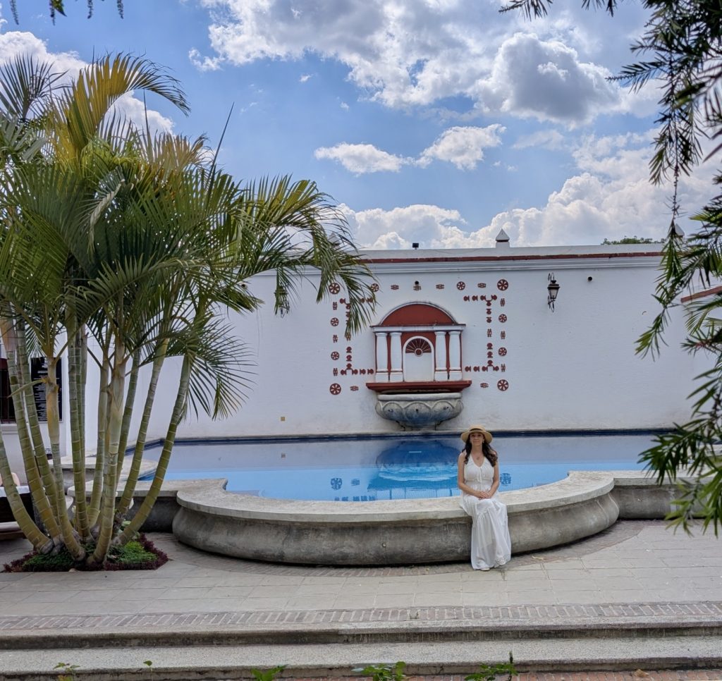  Mujer sentada en el borde de la piscina de Villa Colonial en Antigua Guatemala, rodeada de palmeras bajo un cielo azul con nubes, con la pared ornamental colonial de fondo