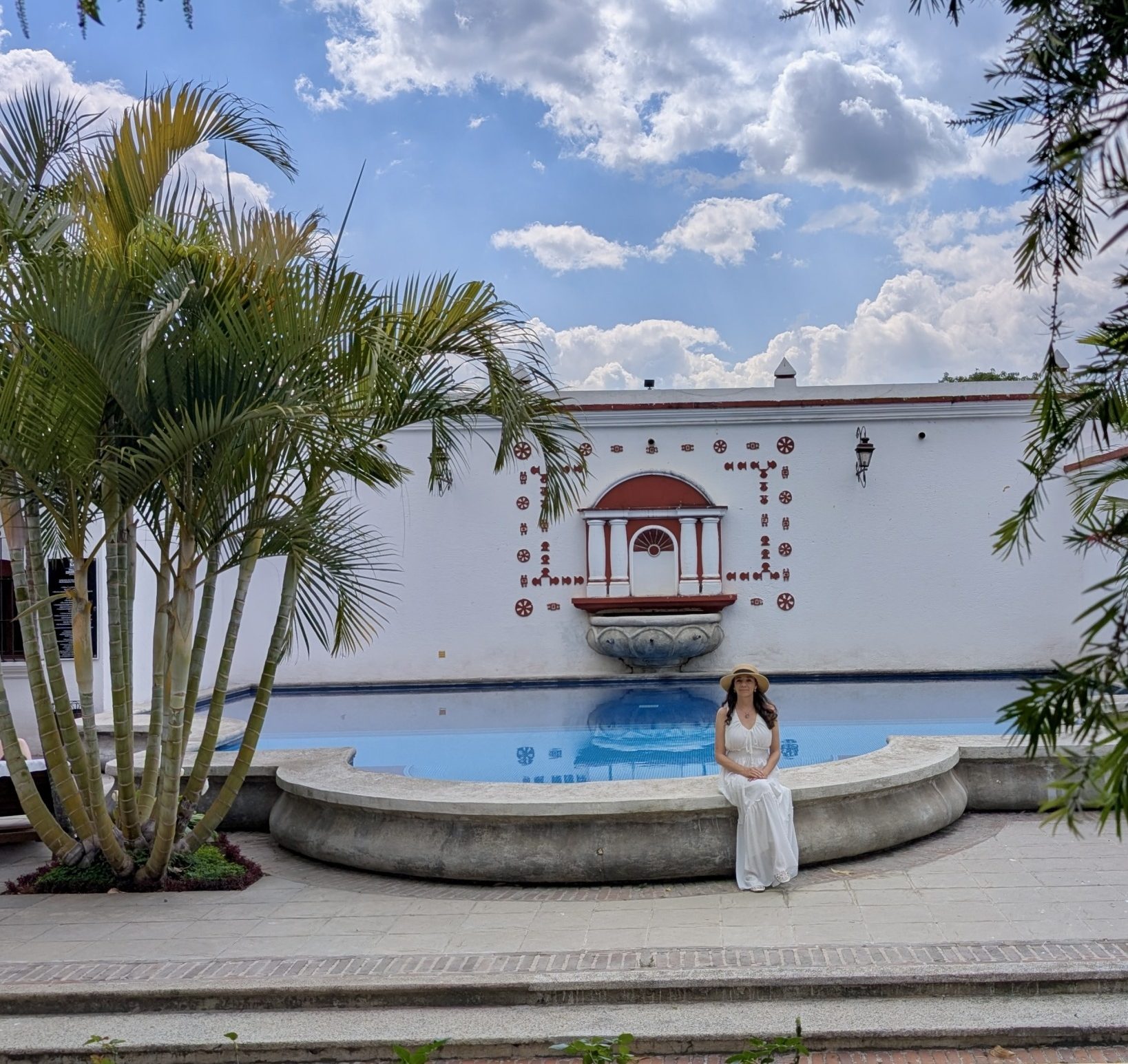 Mujer sentada en el borde de la piscina de Villa Colonial en Antigua Guatemala, rodeada de palmeras bajo un cielo azul con nubes, con la pared ornamental colonial de fondo