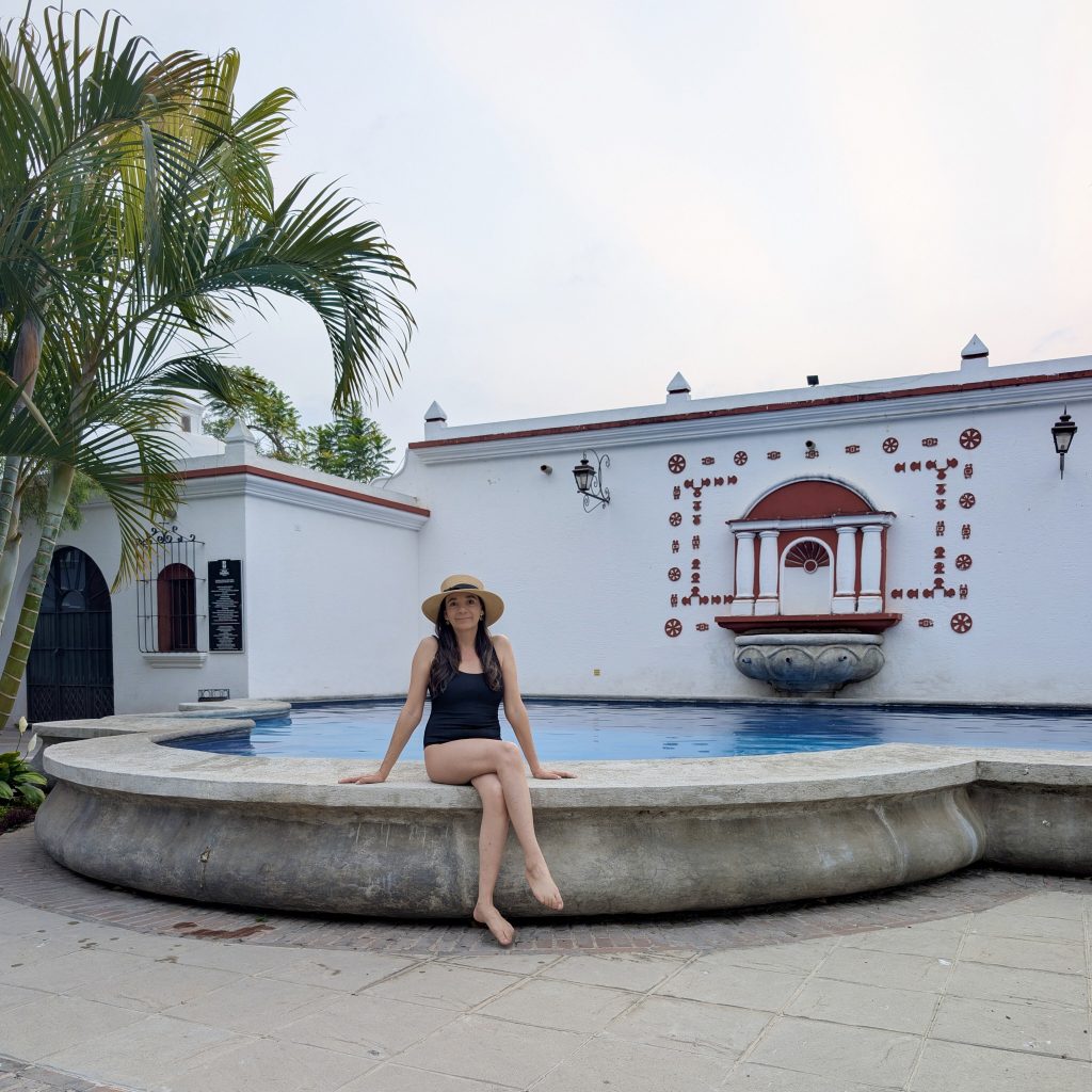 Woman sitting on the stone rim of Villa Colonial's colonial-style pool in Antigua Guatemala, with the ornamental white wall and fountain detail behind her at dusk