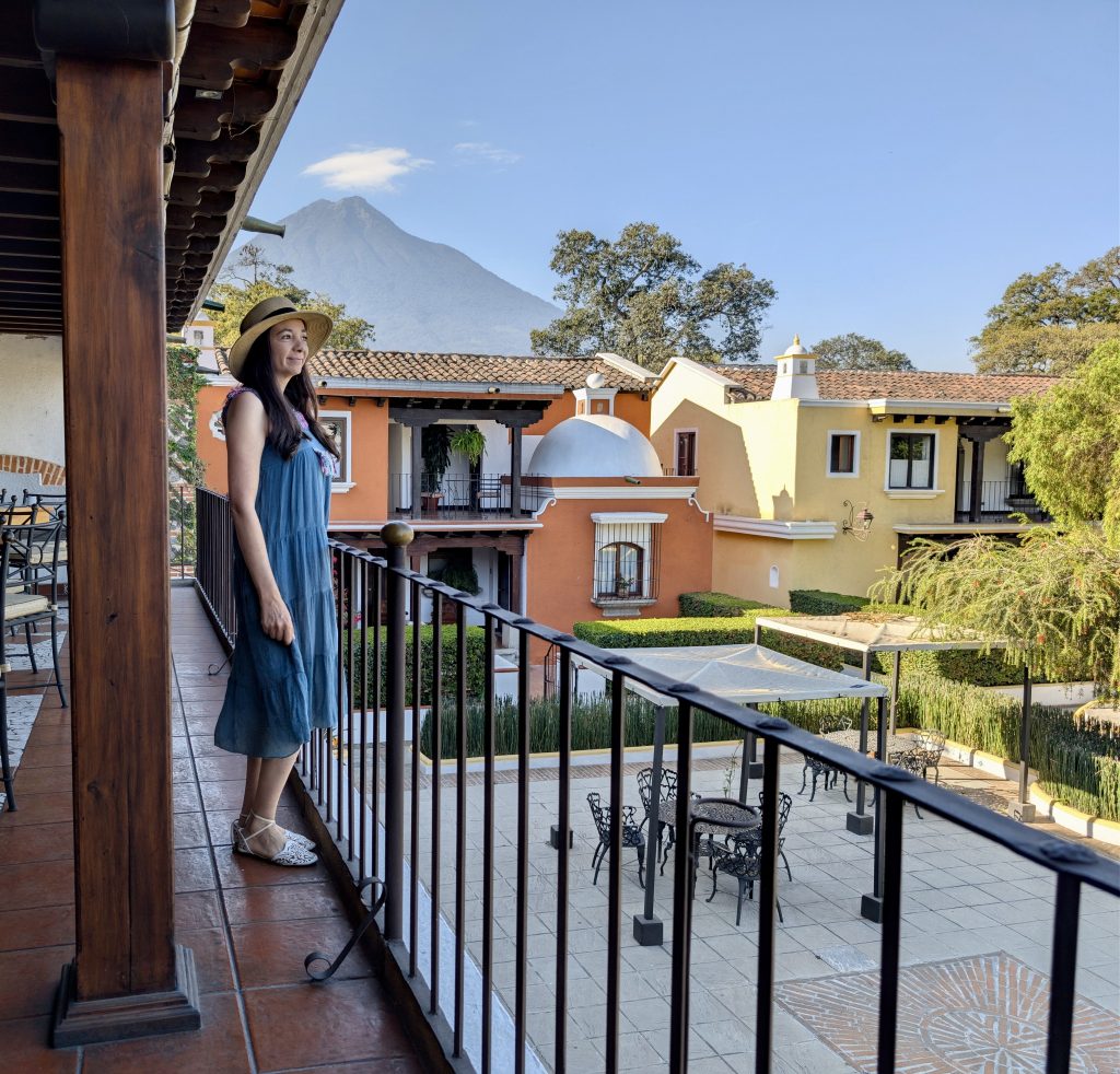 Woman standing on the open-air terrace of Bugambilia restaurant at Villa Colonial in Antigua Guatemala, looking out over the colonial buildings, hotel gardens and Volcán de Agua rising in the background under a clear blue sk