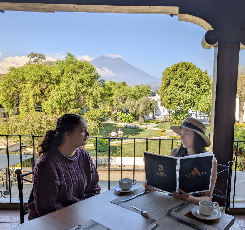 Two guests reviewing the Bugambilia menu at Villa Colonial's open-air restaurant terrace in Antigua Guatemala, with the garden and Volcán de Agua filling the background