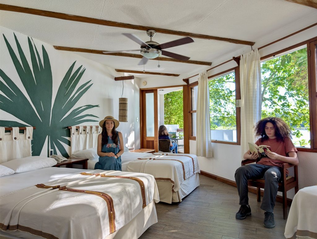 Spacious family room at Villa Maya hotel in Petén Guatemala with two large beds dressed in Guatemalan textile runners, a hand-painted tropical palm leaf wall mural, exposed wood beam ceiling and large windows letting in natural jungle light
