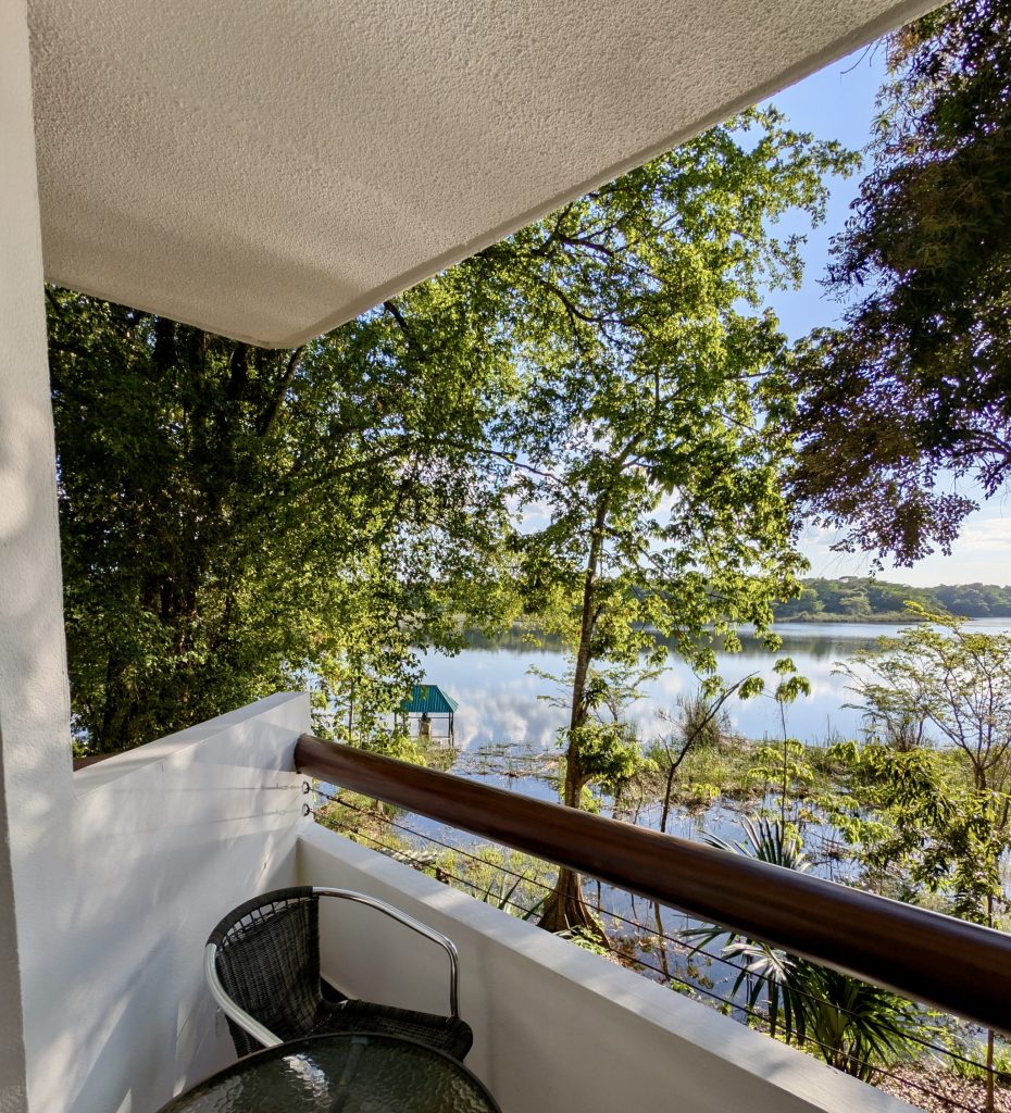 View of the Petenchel lagoon from a second-floor room balcony at Villa Maya hotel in Petén Guatemala, with tropical trees framing calm water reflecting the blue sky