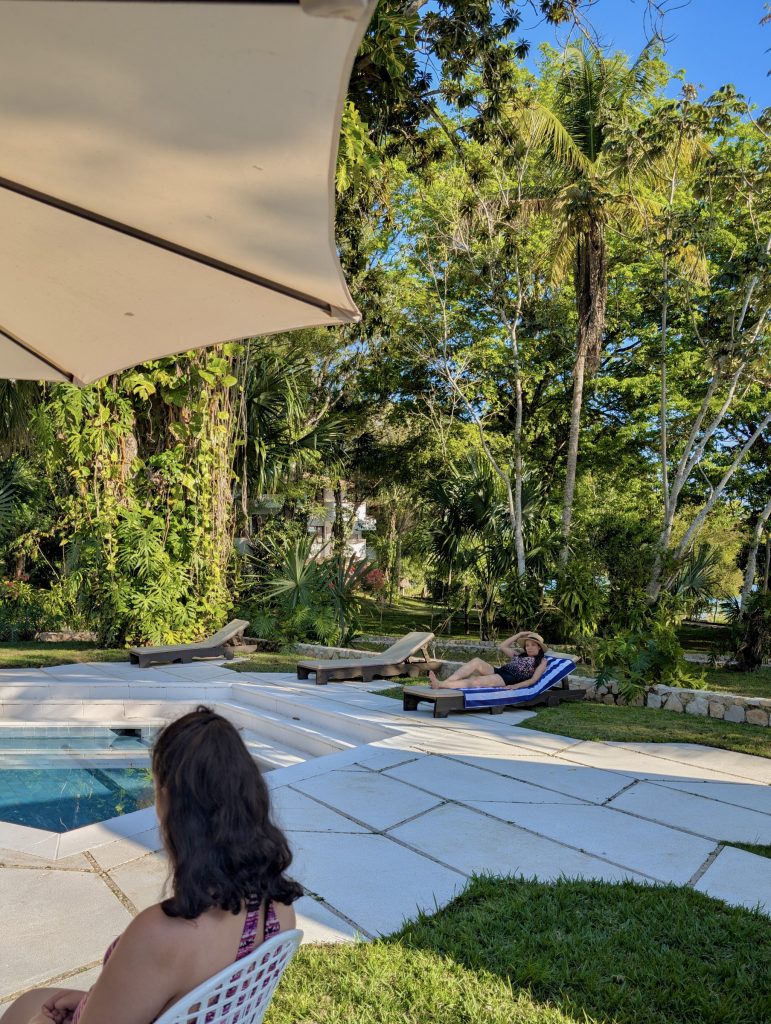 kid's pool area at Villa Maya hotel near Tikal in Petén Guatemala with wooden sun loungers on tropical grass, a large shade umbrella and dense jungle canopy and lagoon visible in the background