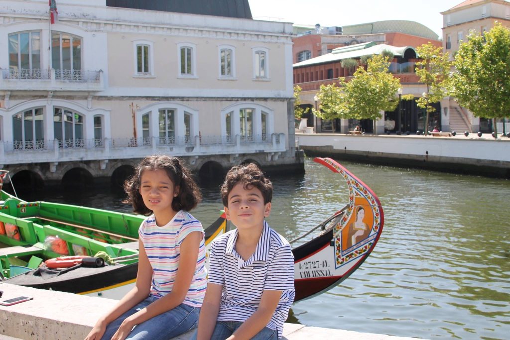 Two children sitting on a canal wall in Aveiro Portugal with colorful moliceiro boat behind them family travel